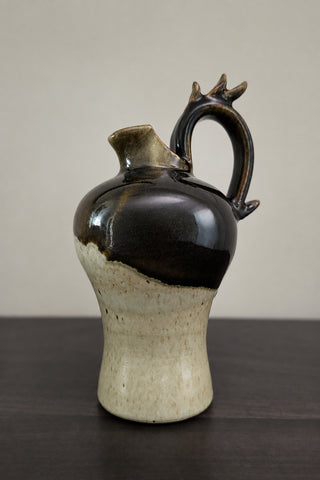 A handmade ceramic pitcher with a glossy dark brown glaze on the upper body, a speckled beige lower body, and a unique antler-like handle, displayed on a dark wooden surface against a plain beige wall.