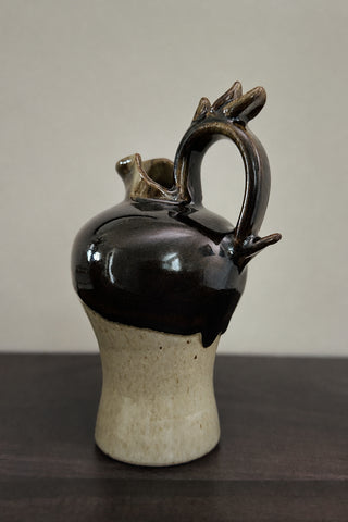 A handmade ceramic pitcher with a glossy black glaze on the upper body and a speckled beige clay base, featuring a unique handle shaped like a small bird and a small pouring spout, set on a dark wooden surface against a neutral light gray background.