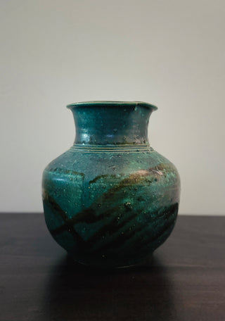 A small ceramic vase with a rounded body and flared neck, glazed in a mottled teal and turquoise finish with dark brown and black brush-like streaks, set on a dark wooden surface against a plain light gray wall.