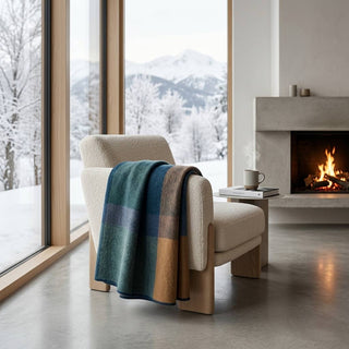 A cream bouclé armchair in a modern living room, draped with a teal, blue, and tan plaid throw blanket. Large windows reveal a snowy mountain landscape, and a concrete fireplace with a fire burns in the background.