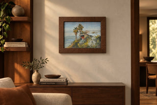 A framed landscape painting of a coastal cottage scene hangs on a warm beige wall in a styled interior room with wooden furniture and decor.