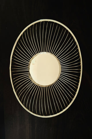 An oval basket with a gold-toned frame and a dense ring of thin radial metal wires, shown against a dark background. The reflective center shows a distorted reflection of a person taking the photo.