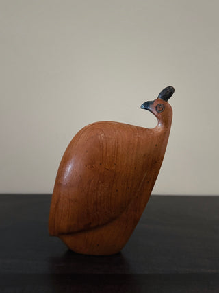 A carved wooden bird sculpture, resembling a peacock or quail, with a smooth reddish-brown wood finish and a small dark blue accent on its head, standing on a dark surface against a plain beige background.