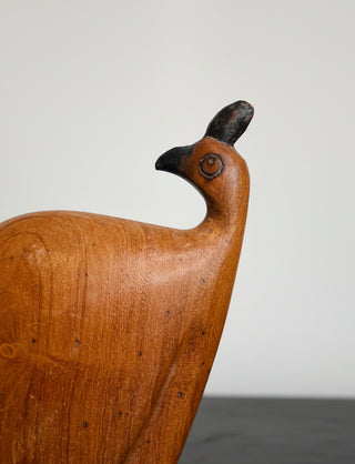 A close-up of a carved wooden bird figurine, likely a quail or similar small bird, with a smooth warm brown body and a dark beak and crest, set against a plain light background.