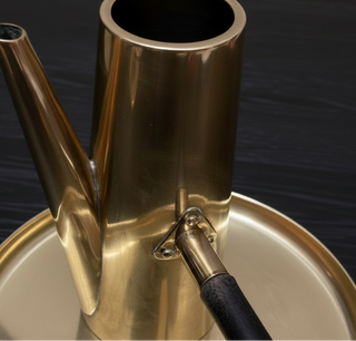 A close-up of a polished brass or bronze metal vessel with a spout and a small black-handled tool resting against the base, set on a wooden surface with a neutral wall in the background.