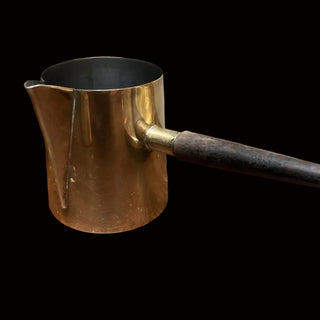 A small brass coffee pour-over pot with a dark wooden handle, sitting on a scratched brown wooden table against a plain beige wall.