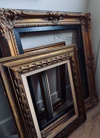 Three ornate picture frames in different sizes, including two gold frames and one black frame, leaning against a light gray wall on a wooden floor.
