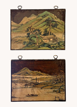 Two vintage Japanese-style landscape wall hangings displayed side-by-side on a white wall. The top hanging depicts a green mountain village scene, and the bottom hanging depicts a river or harbor scene with a boat and mountains.