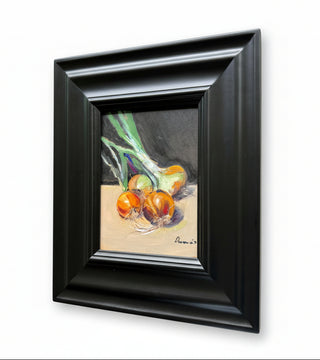 A framed still life painting of onions hanging on a light-colored wall. The artwork features three orange onions with green tops on a beige surface against a dark gray background, enclosed in a thick black frame.