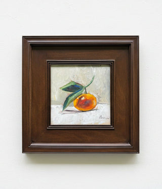 A framed still life painting of an orange with green leaves, presented in a dark brown wooden frame against a light gray wall.