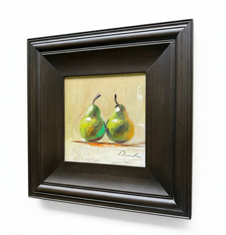 A framed still life painting of two green pears on a cream background, housed in a dark brown wooden frame, shown hanging on a light-colored wall.