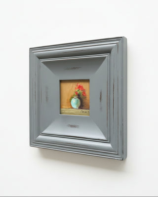 A square silver-gray distressed picture frame holding a small still life painting of a green vase with red flowers, mounted on a light beige wall.