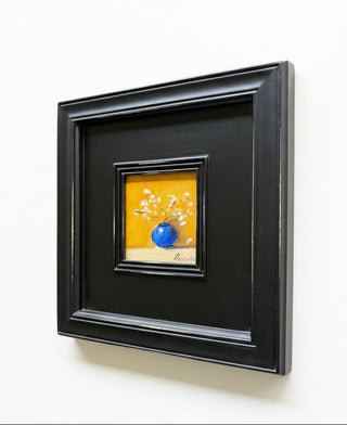 A square black picture frame containing a small colorful painting of white flowers and a blue sphere on a yellow background, mounted on a light beige wall.