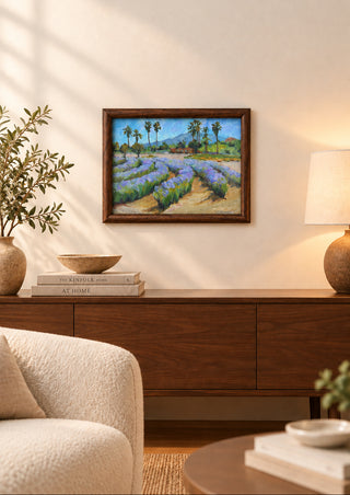 A framed landscape painting of a lavender field with purple flowers, green foliage, and palm trees under a blue sky, set in a dark wood frame on a light gray surface.