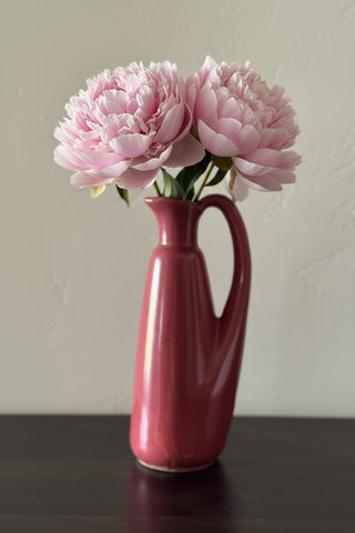 A glossy, mid-century modern ceramic pitcher in a dusty rose or muted terracotta pink, standing on a dark wooden surface against a textured off-white wall.