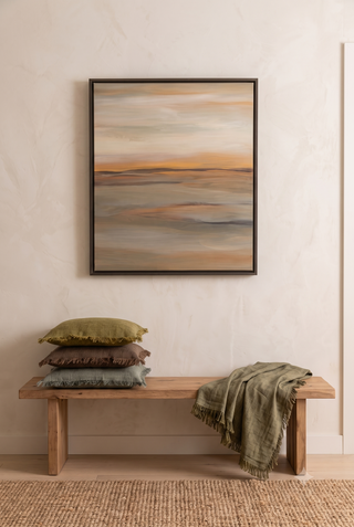 A framed abstract landscape painting hangs on a textured beige wall above a wooden bench with stacked pillows and a draped throw blanket.