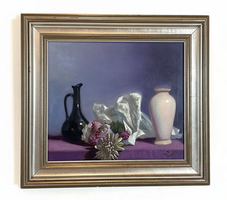 A framed still life oil painting depicting a dark glass pitcher, a white vase, and flowers on a purple tablecloth against a dark blue background, enclosed in a distressed silver frame.