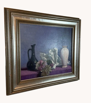 A framed still life painting featuring a dark jug, white draped fabric, and a white vase on a purple surface against a dark blue background, housed in a wide ornate silver frame and mounted on a light wall.